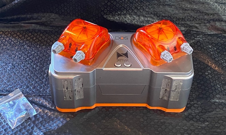 May include: A gray and orange Neptune Systems DOS pump with two orange dosing heads. The pump has a label that says "DOS" and "Neptune Systems".
