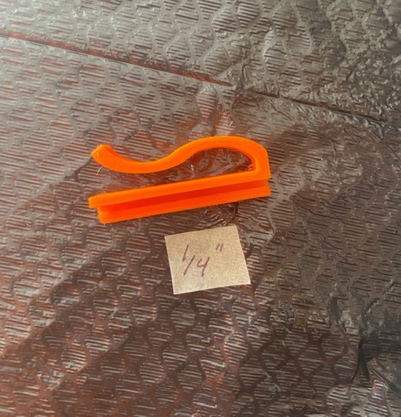 May include: Orange plastic clip measuring 1/4 inch. The clip is shaped like a curved line with a flat base.
