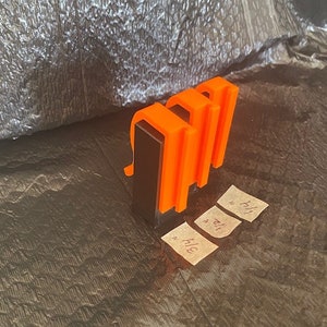 May include: Three orange plastic clips attached to a black rectangular base. The clips are designed to hold papers or other items together.