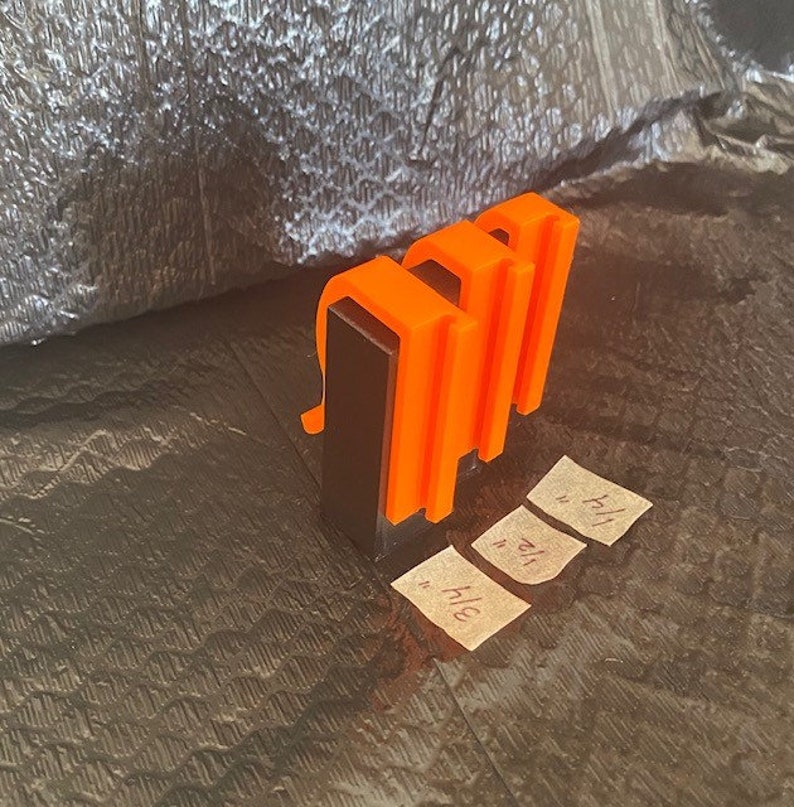 May include: Three orange plastic clips attached to a black rectangular base. The clips are designed to hold papers or other items together.
