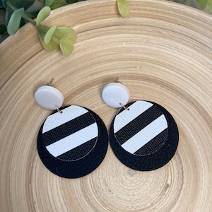 May include: A pair of black and white striped circle earrings with a white circle on top. The earrings are on a wooden surface.