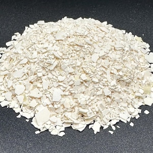May include: A pile of off-white, irregularly shaped fragments. The pieces vary in size and have a slightly textured appearance. The fragments are scattered on a dark surface, creating a contrast that highlights their form.