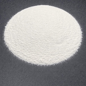 May include: A circular pile of fine, white powder against a dark gray background. The powder has a slightly textured surface, creating subtle shadows and highlights. The overall shape is a rounded mound.