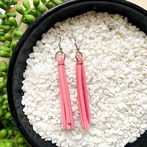 May include: A pair of pink suede tassel earrings with silver-colored ear wires. The earrings are displayed in a black bowl filled with white pebbles, with a wooden slice and green plant in the background.