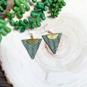May include: A pair of handmade earrings featuring triangular, sage green leather pendants with a woven texture. Each earring has a gold-colored, half-circle accent and gold-colored ear hooks. The earrings are displayed on a light wood surface with green plant accents.