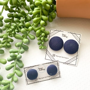 May include: Navy blue fabric-covered button earrings on white cards. The cards have a geometric border and the text "Ginger Darlings Co" with an Instagram handle. Green succulent plant and a terracotta pot are in the background.