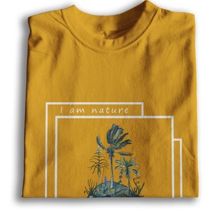 Puede incluir: Una camiseta amarilla mostaza con un estampado gr&aacute;fico blanco de una escena tropical con el texto "I am nature" en un cuadro blanco.