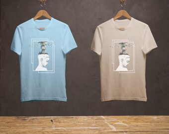 Puede incluir: Dos camisetas con un diseño gráfico de una cabeza con un paisaje en la parte superior. La camiseta de la izquierda es azul claro y la camiseta de la derecha es beige. El diseño gráfico presenta una cabeza con un paisaje en la parte superior, incluyendo árboles, edificios y pájaros. El texto "Dream Believer" está impreso en el diseño gráfico.