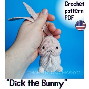 Könnte beinhalten: Ein gehäkelter rosa Hase mit schwarzen Augen und einem gestickten Lächeln. Der Hase wird von einer Hand gehalten und der Text "Dick the Bunny" steht unter dem Bild.