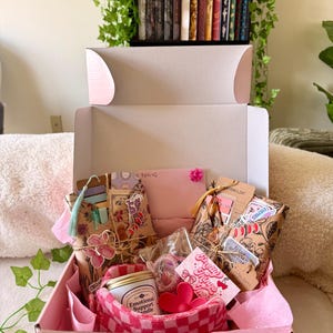 Puede incluir: Una caja de regalo rosa llena de varios artículos, incluyendo una vela, etiquetas decorativas y paquetes envueltos. La caja contiene una cesta a cuadros rosa y roja, un objeto en forma de corazón rosa y una vela "Emotional Support Candle".