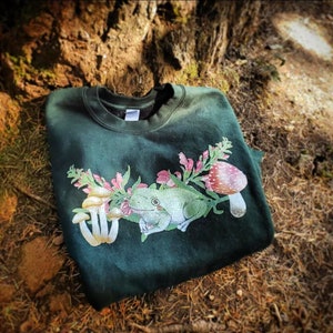May include: A dark green sweatshirt with a white frog, pink flowers, and mushrooms printed on the front.