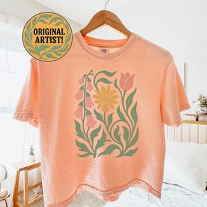 May include: A peach-colored short-sleeve t-shirt featuring a central graphic print of a yellow daisy, pink bell-shaped flowers, and a pink tulip, all surrounded by green leafy stems. The shirt has a crew neckline and is displayed on a wooden hanger.