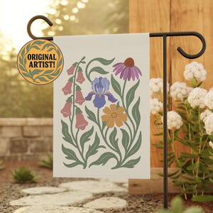 May include: A cream-colored garden flag featuring a floral design with pink, purple, blue, and yellow flowers and green leaves. The flag is displayed on a black metal stand. The flag also has a circular emblem that says "ORIGINAL ARTIST!"