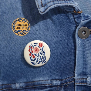 May include: Two round buttons are pinned to a denim jacket. One button has the text "ORIGINAL ARTIST!" in a yellow and blue design. The other button features a floral design with red, pink, and blue elements.