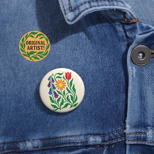 May include: Two round buttons pinned to a denim jacket. One button features the text "ORIGINAL ARTIST!" in a green and yellow design. The other button displays a floral design with a red tulip, orange flower, and purple and blue bell-shaped flowers.