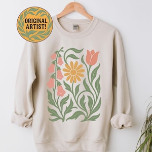 May include: Cream-colored sweatshirt featuring a floral design with a yellow daisy, pink bell-shaped flowers, and a peach tulip. The design is framed by green leaves and stems. The text "ORIGINAL ARTIST!" is in a circular emblem.
