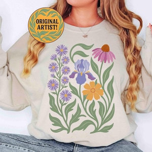 May include: Beige sweatshirt featuring a floral design with purple, yellow, and pink flowers, green leaves, and stems. The sweatshirt also has the text "ORIGINAL ARTIST!" in a circular design.