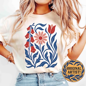 May include: Cream-colored t-shirt featuring a floral design with red bell-shaped flowers, a pink daisy, and a red tulip, all set against blue leaves and stems. The shirt also has the text "ORIGINAL ARTIST!" in a circular design.