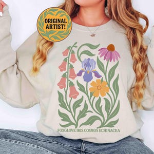May include: Beige sweatshirt featuring a floral design with foxglove, iris, cosmos, and echinacea. The design includes pink, purple, yellow, and green flowers with the text "FOXGLOVE IRIS COSMOS ECHINACEA" at the bottom. A circular "ORIGINAL ARTIST!" logo is at the top.