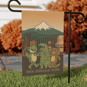 May include: A garden flag featuring a cartoon illustration of a frog, dinosaur, and teddy bear in front of a city skyline and mountain. The flag has the text "FROG DINOSAUR TEDDY" at the bottom.