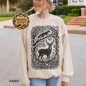 May include: A sand-colored crewneck sweatshirt featuring a black graphic print of a deer, a fox, and a turtle surrounded by forest foliage and mushrooms. A circular badge on the upper left reads ORIGINAL ARTIST! in black text on a yellow background.