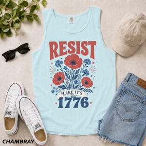 May include: A light blue tank top featuring a red and blue floral graphic with the text RESIST LIKE IT'S 1776. The design includes red poppies and blue cornflowers arranged in a bouquet, with the year 1776 printed in large blue block lettering.