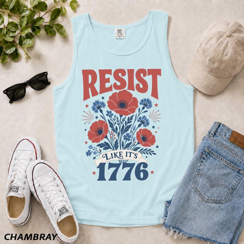 May include: A light blue tank top featuring a red and blue floral graphic with the text RESIST LIKE IT'S 1776. The design includes red poppies and blue cornflowers arranged in a bouquet, with the year 1776 printed in large blue block lettering.