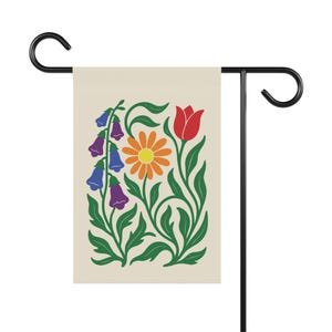 May include: A garden flag featuring a floral design with a cream background. The design includes a red tulip, an orange daisy, and purple and blue bell-shaped flowers, all with green stems and leaves. The flag is mounted on a black metal stand.
