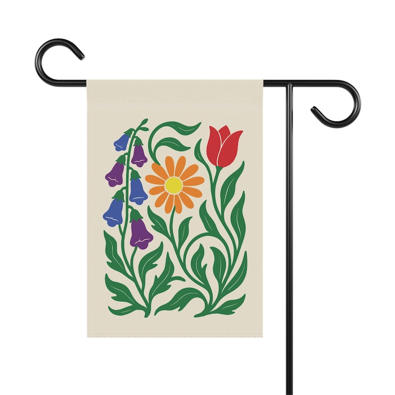 May include: A garden flag featuring a floral design with a cream background. The design includes a red tulip, an orange daisy, and purple and blue bell-shaped flowers, all with green stems and leaves. The flag is mounted on a black metal stand.