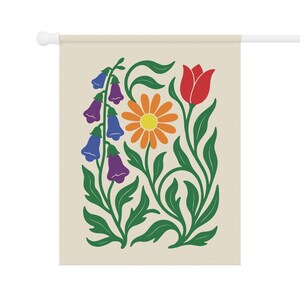 May include: A beige garden flag featuring a colorful floral design. The design includes a red tulip, an orange daisy, and blue and purple bell-shaped flowers, all with green leaves and stems. The flag is mounted on a white pole.