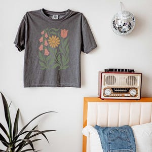 May include: A charcoal gray short-sleeve t-shirt featuring a central graphic print of a yellow daisy, a pink tulip, and pink bell-shaped flowers surrounded by green leafy stems and foliage. The shirt has a crew neckline and a relaxed fit.