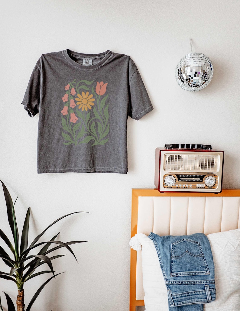 May include: A charcoal gray short-sleeve t-shirt featuring a central graphic print of a yellow daisy, a pink tulip, and pink bell-shaped flowers surrounded by green leafy stems and foliage. The shirt has a crew neckline and a relaxed fit.