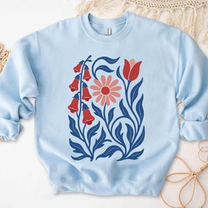 May include: Light blue sweatshirt featuring a floral design. The design includes red bell-shaped flowers, a pink daisy, and blue leaves and stems. The sweatshirt has a crew neck and long sleeves with ribbed cuffs.