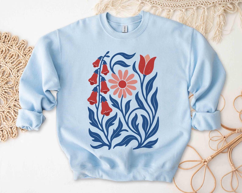 May include: Light blue sweatshirt featuring a floral design. The design includes red bell-shaped flowers, a pink daisy, and blue leaves and stems. The sweatshirt has a crew neck and long sleeves with ribbed cuffs.