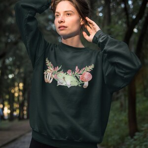 May include: A dark green sweatshirt with a watercolor illustration of a green frog sitting on a branch with pink flowers and red mushrooms.