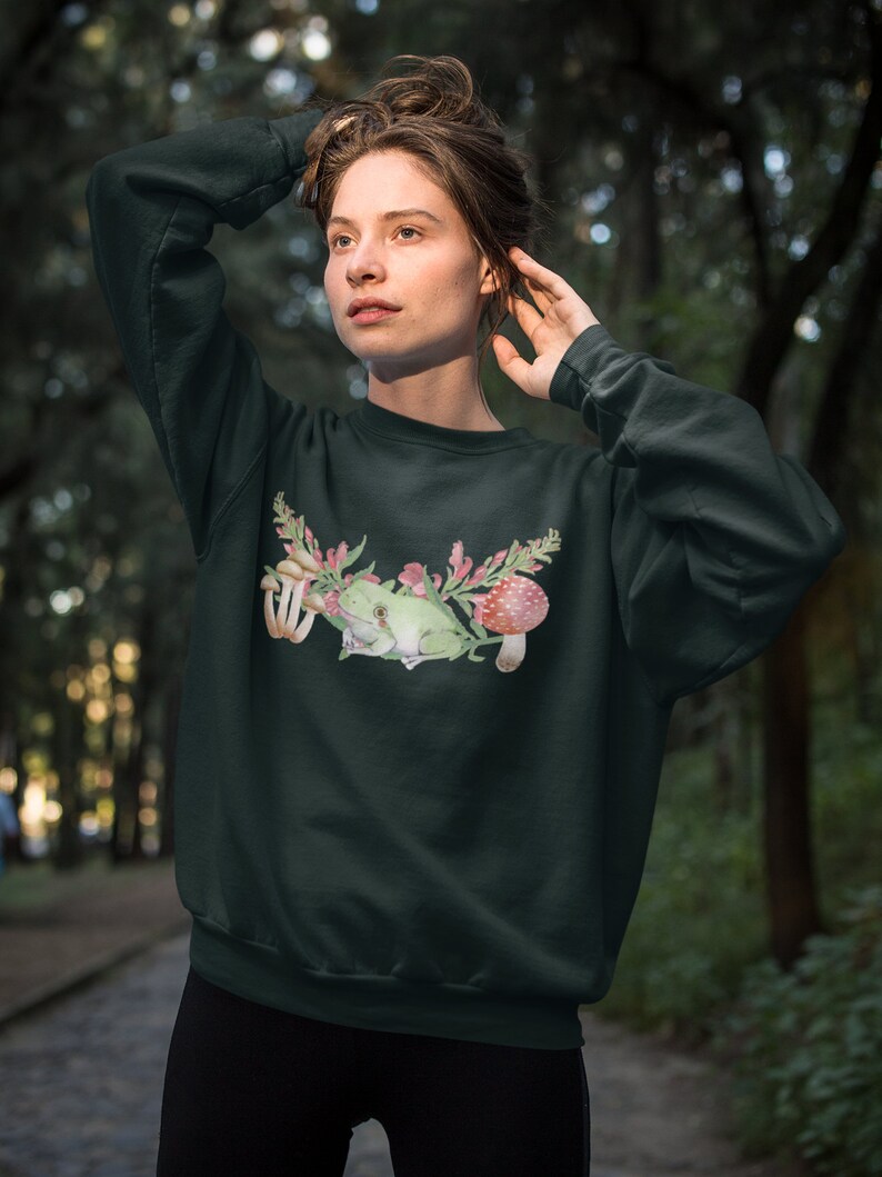 May include: A dark green sweatshirt with a watercolor illustration of a green frog sitting on a branch with pink flowers and red mushrooms.