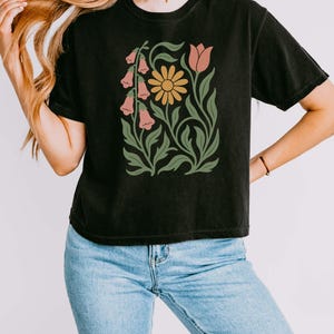 May include: A black short-sleeve t-shirt featuring a central graphic print of a yellow daisy, a pink tulip, and pink bell-shaped flowers with green stems and leaves. The shirt is paired with light blue denim jeans.