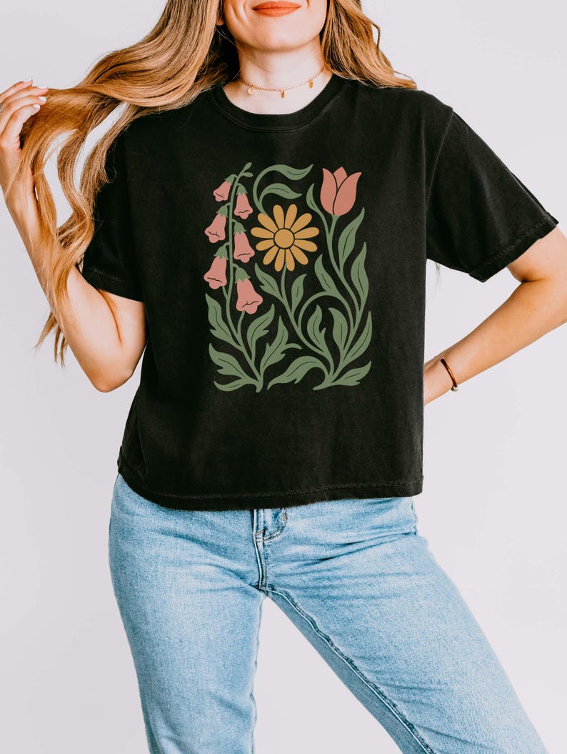 May include: A black short-sleeve t-shirt featuring a central graphic print of a yellow daisy, a pink tulip, and pink bell-shaped flowers with green stems and leaves. The shirt is paired with light blue denim jeans.