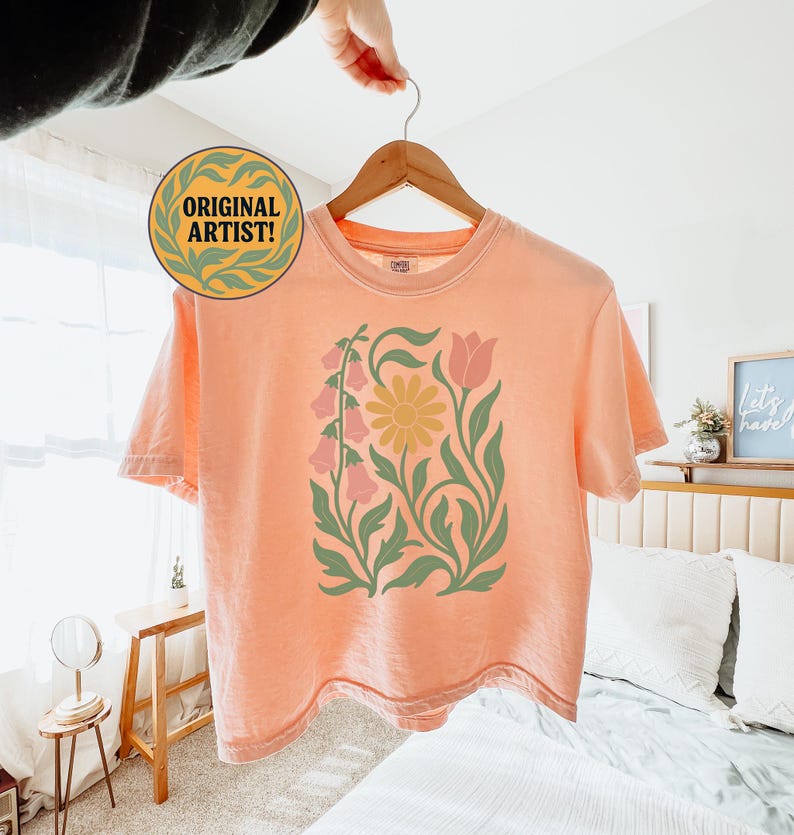 May include: A peach-colored short-sleeve t-shirt featuring a central graphic print of a yellow daisy, pink bell-shaped flowers, and a pink tulip, all surrounded by green leafy stems. The shirt has a crew neckline and is displayed on a wooden hanger.