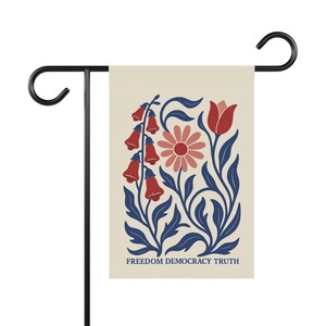 Peut inclure: Un drapeau de jardin rectangulaire suspendu &agrave; un support en m&eacute;tal noir. Le drapeau est de couleur cr&egrave;me avec une illustration botanique de fleurs rouges et roses et de feuilles bleu fonc&eacute;. Le texte FREEDOM DEMOCRACY TRUTH est imprim&eacute; en bleu.