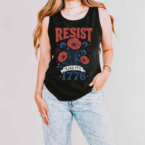 May include: A black sleeveless tank top featuring a graphic print with the red text RESIST above a floral illustration of red poppies and blue cornflowers. Below the flowers is a white banner with the blue text LIKE IT'S 1776 and small stars.