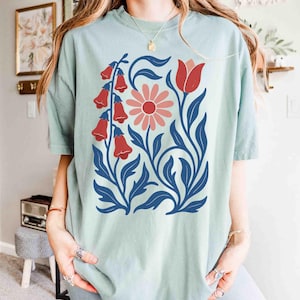 May include: A light green t-shirt featuring a floral design. The design includes red bell-shaped flowers, a pink daisy, and blue leaves and stems. The shirt has short sleeves and a relaxed fit, suitable for casual wear.