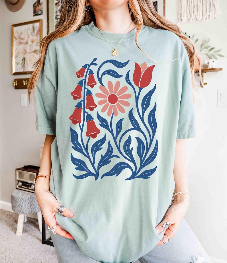 May include: A light green t-shirt featuring a floral design. The design includes red bell-shaped flowers, a pink daisy, and blue leaves and stems. The shirt has short sleeves and a relaxed fit, suitable for casual wear.