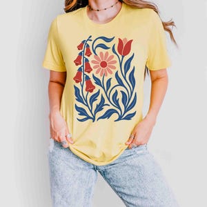 May include: Yellow t-shirt featuring a floral design. The graphic includes red bell-shaped flowers, a pink daisy, and blue leaves and stems. The shirt is a short-sleeved crew neck.