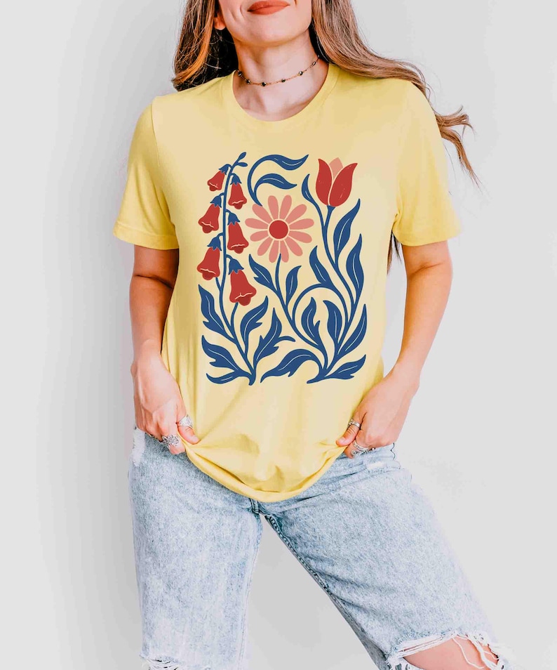 May include: Yellow t-shirt featuring a floral design. The graphic includes red bell-shaped flowers, a pink daisy, and blue leaves and stems. The shirt is a short-sleeved crew neck.