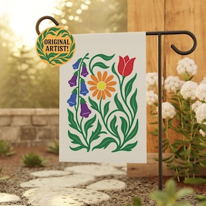 May include: A cream-colored garden flag featuring a floral design with a red tulip, orange daisy, and purple and blue bellflowers. The flag is displayed on a black metal stand. The flag has the text "ORIGINAL ARTIST!" on a circular tag.