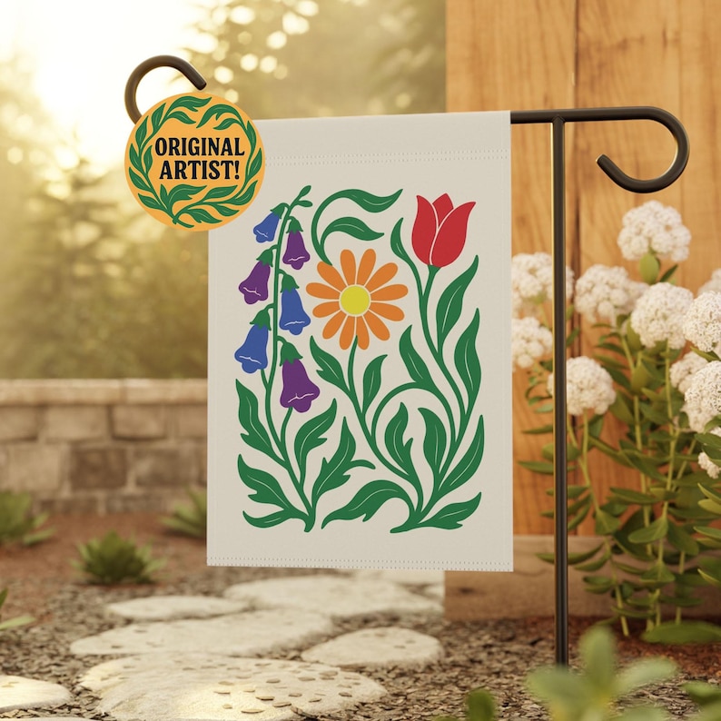 May include: A cream-colored garden flag featuring a floral design with a red tulip, orange daisy, and purple and blue bellflowers. The flag is displayed on a black metal stand. The flag has the text "ORIGINAL ARTIST!" on a circular tag.