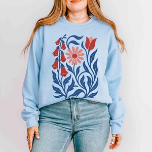 May include: Light blue sweatshirt featuring a floral design. The graphic includes red bell-shaped flowers, a pink daisy, and blue leaves and stems. The sweatshirt is a crew neck style.