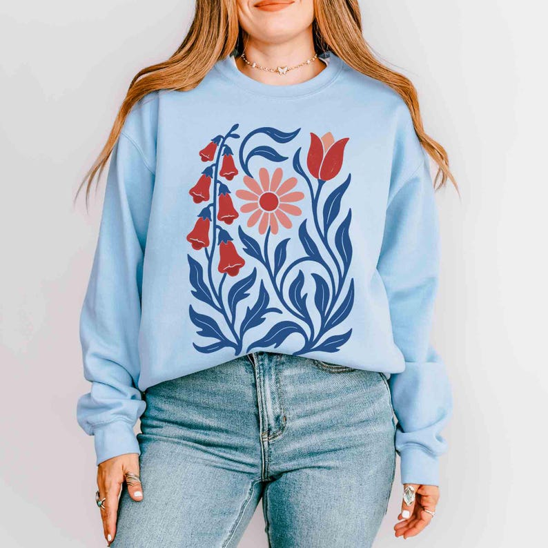 May include: Light blue sweatshirt featuring a floral design. The graphic includes red bell-shaped flowers, a pink daisy, and blue leaves and stems. The sweatshirt is a crew neck style.