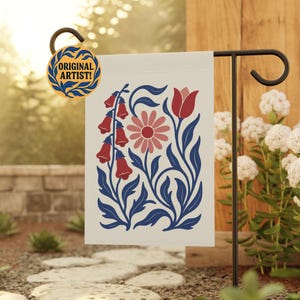 May include: A garden flag featuring a floral design with red, pink, and blue elements. The flag displays a stylized flower and leaf pattern on a cream background. The words "ORIGINAL ARTIST!" are on a circular tag.